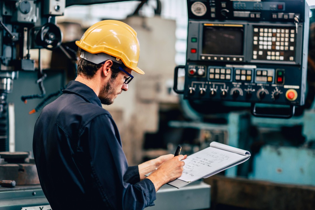 A factory worker wearing a yellow hard hat and safety glasses writes on a clipboard while inspecting industrial machinery in a manufacturing environment. The background shows a control panel and production equipment.