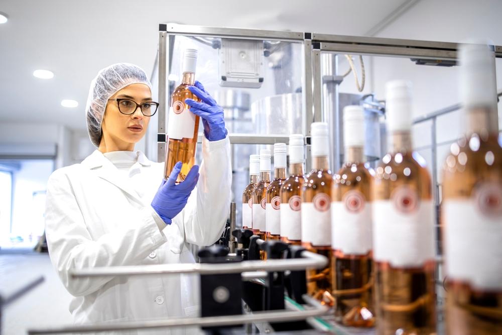 A women standing beside a conveyor of wine bottles and she has one bottle in her hands inspecting the bottle for imperfections.